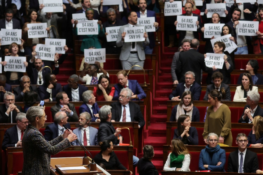 Parliamentarians stand, holding printed signs that read '64 anon, cest non'. A woman looks at them from a lectern.