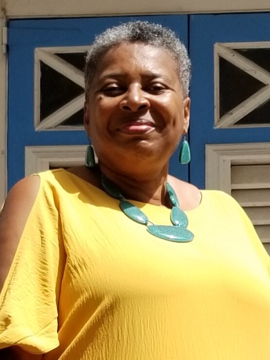 A middle-aged woman with African-Caribbean heritage wearing a yellow top and a green necklace smiles