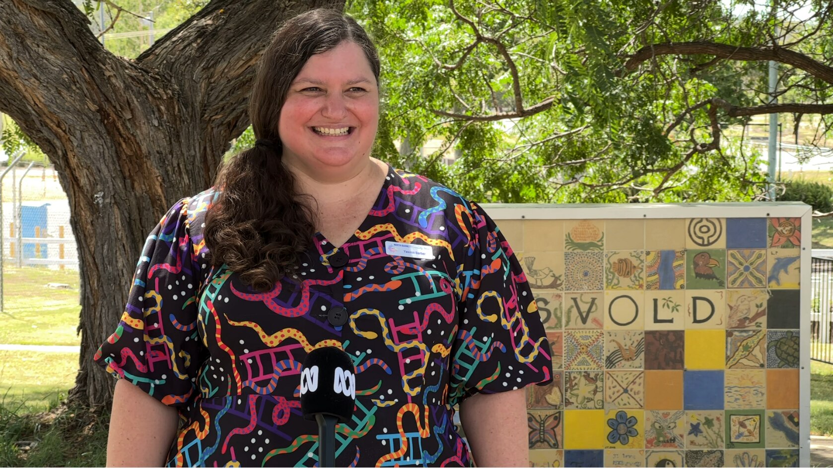 A woman in a dress with squiggles of pink, yellow and green stands in a park in front of a tree and a sign that reads Eidsvold. 