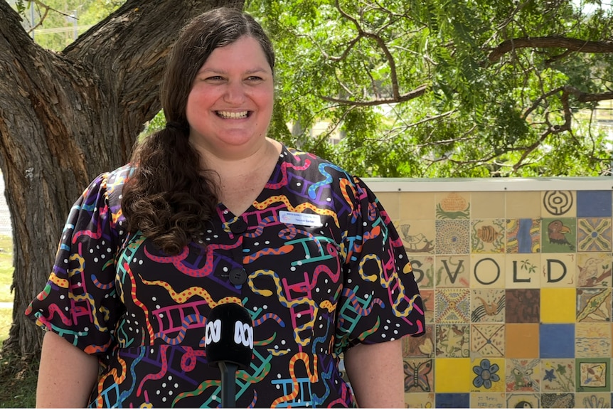 A woman in a dress with squiggles of pink, yellow and green stands in a park in front of a tree and a sign that reads Eidsvold. 