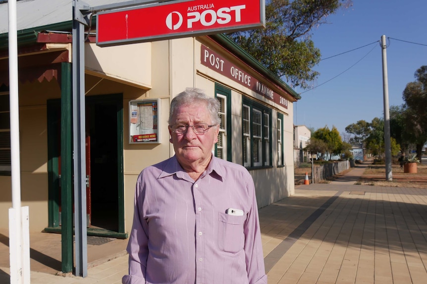 An elderly man stands outside an old-style cream-coloured post office underneath an Australia Post sign.