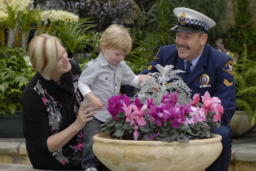 A man in a police uniform and a woman smiling with a small child in front of flowers,
