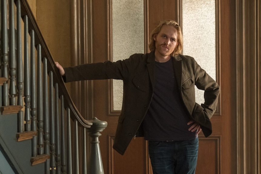 Film still of a bearded Wyatt Russell as David leaning against a staircase railing in The Woman in the Window