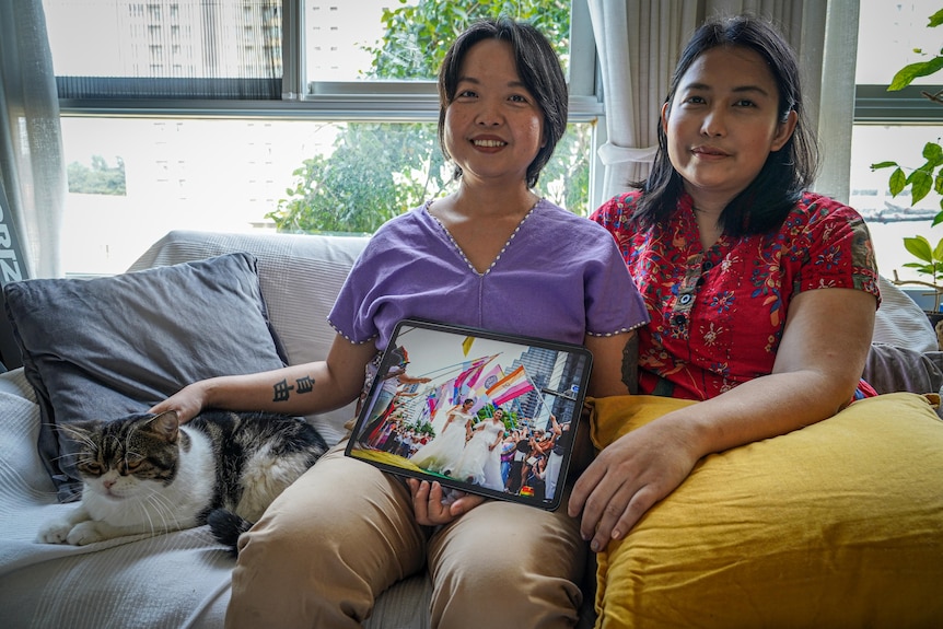 Two women sitting on a couch holding a photo and petting a cat 