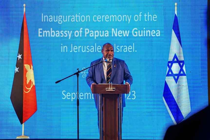 Man stands on podium between PNG and Israel flags in front of blue background. 