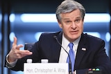 A man in a navy suit gestures with his right hand as he speaks into a desk mounted microphone.