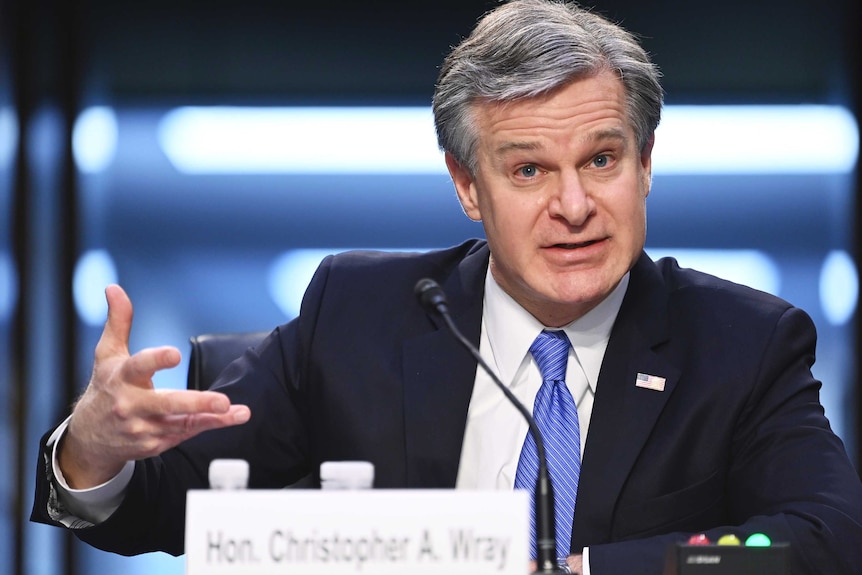 A man in a navy suit gestures with his right hand as he speaks into a desk mounted microphone.