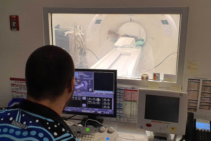 view of radiologists computer screen with CT scanner in background through glass. Piece of wood in CT