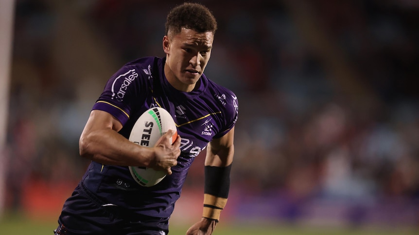 A Melbourne Storm NRL player runs the ball tucked under his right arm.