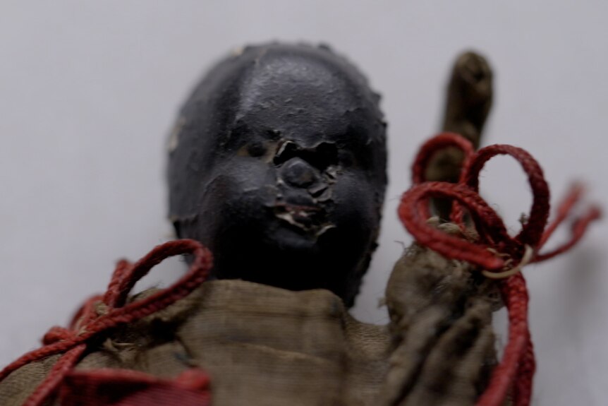 A front on closeup of a very old black doll with no hair. It wears a brown dress with red ribbons tied on its shoulders