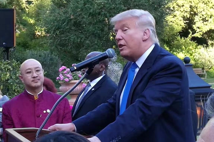 Donald Trump at lectern wearing suit speaks into microphone as man in background watches