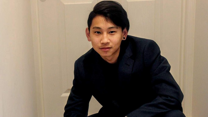 Dillon Wu, wearing a dark blue suit with flicked black hair and a black earing, crouches in front of a white door in a hallway.