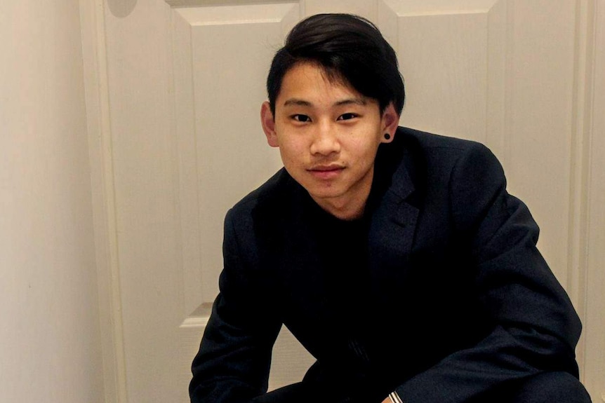 Dillon Wu, dressed in a dark blue suit with tousled black hair and a black ear, crouches in front of a white door in a hallway.
