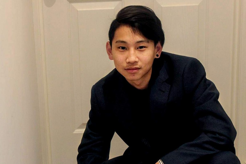 Dillon Wu, wearing a dark blue suit with flicked black hair and a black earing, crouches in front of a white door in a hallway.