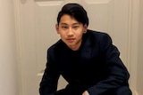 Dillon Wu, wearing a dark blue suit with flicked black hair and a black earing, crouches in front of a white door in a hallway.