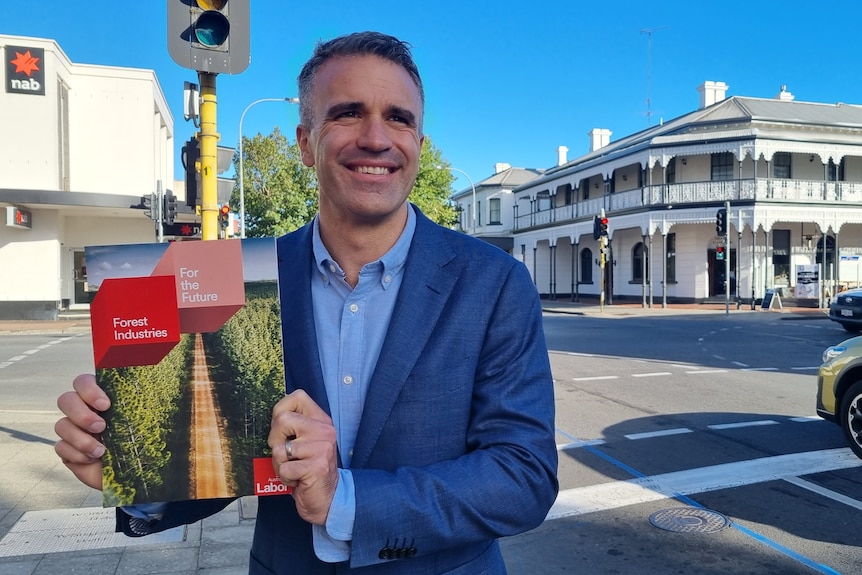 A man in a suit jacket smiles at the camera holding up a forestry booklet