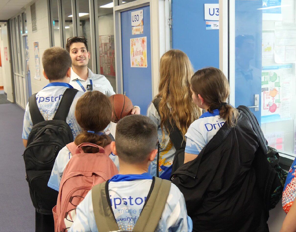 A smiling male teacher in a school hallway holds a basketball while facing 5 students with backpacks and blue-and-white uniforms
