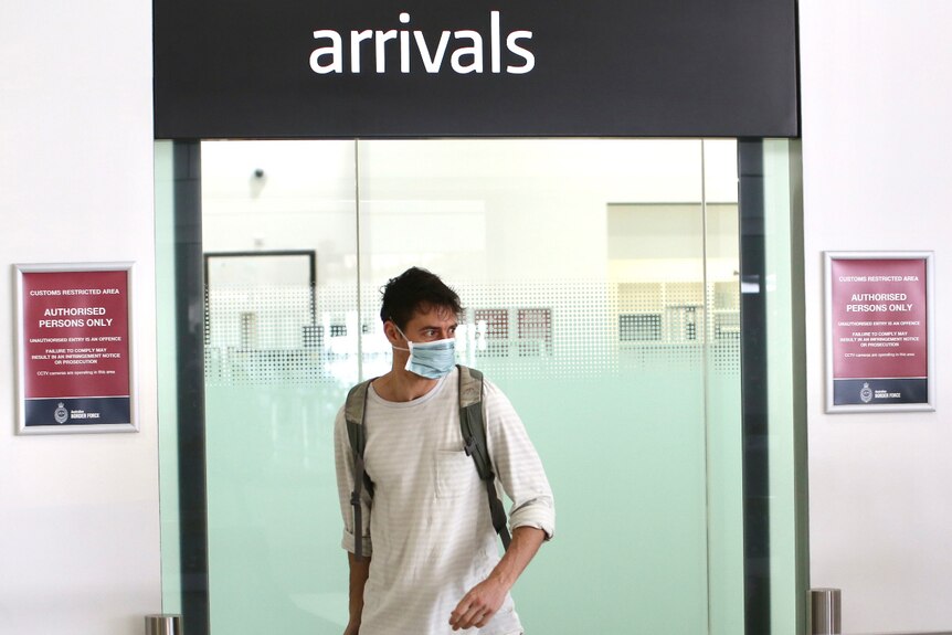 A young man wearing a blue face mask walks through the arrival gate at Perth Airport with luggage