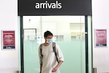 A young man wearing a blue face mask walks through the arrival gate at Perth Airport with luggage