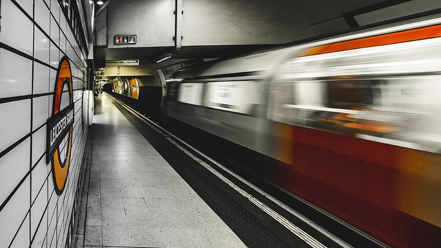 A london tube train speeds by blurred in past a sign for Leicester Square in tones of orange and grey.