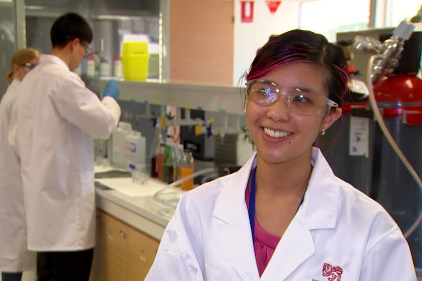 A woman wearing safety glasses and a white coat smiles at the camera.
