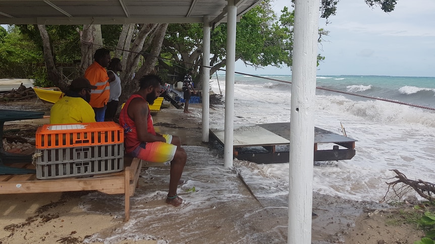 A group of Torres Strait Islander men watch as rising waters lap at their feet.