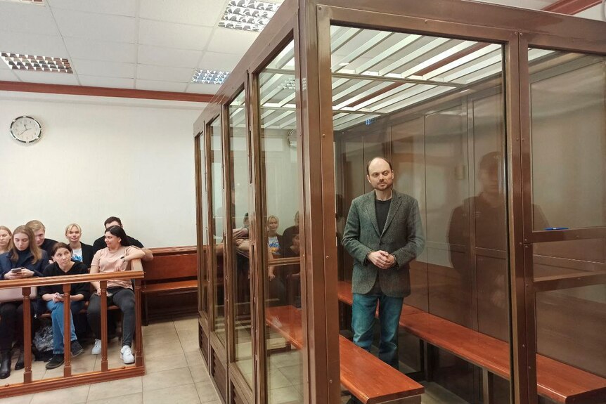 A man stands behind glass in the dock in a court room while people can be seen sitting on a bench.