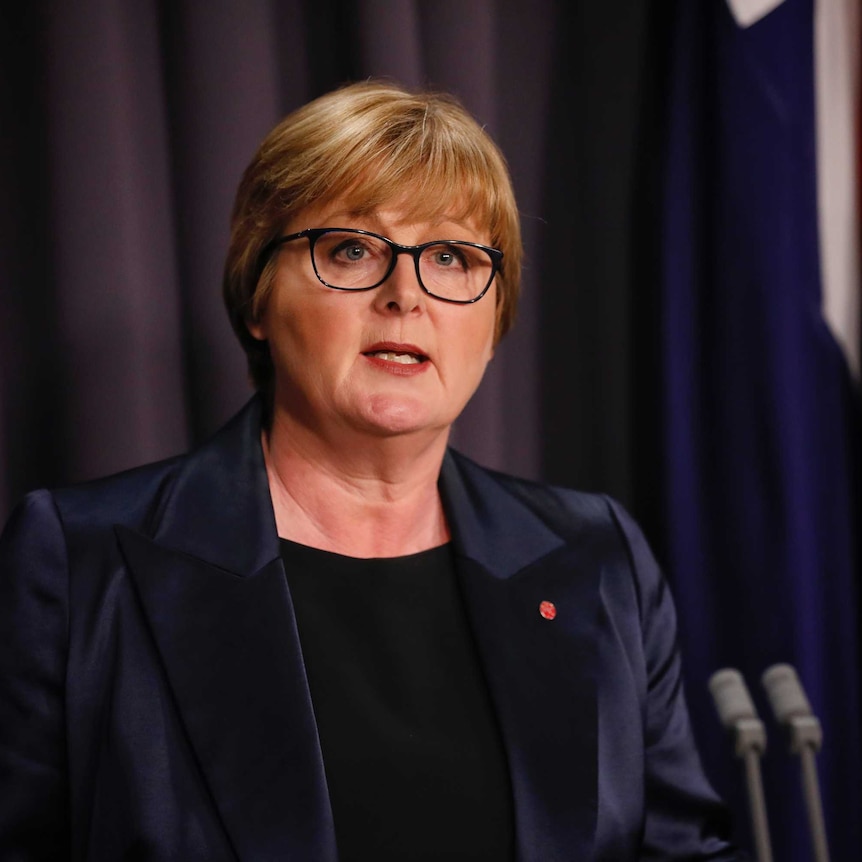 A woman with short blonde hair and glasses speaking a lecturn in front of an australian flag