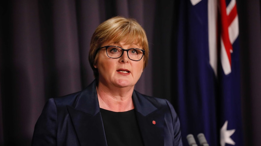 A woman with short blonde hair and glasses speaking a lecturn in front of an australian flag