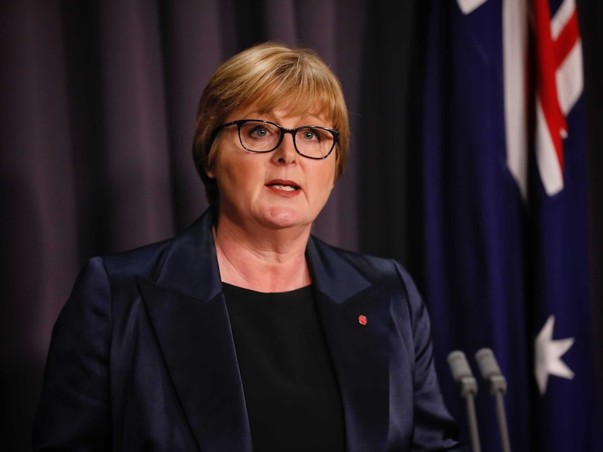 A woman with short blonde hair and glasses speaking a lecturn in front of an australian flag