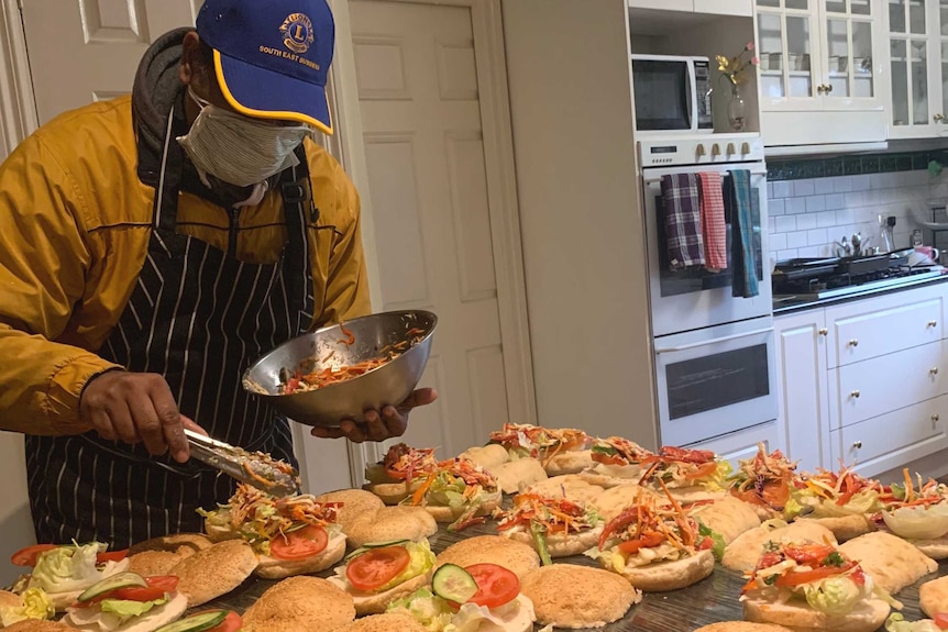 Daman uses tongs to put filling on burgers on a bench in his home kitchen.