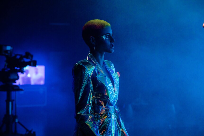 A woman walks to the right of the frame bathed in blue light. She wears an iridescent trench, her hair is dyed multiple colours