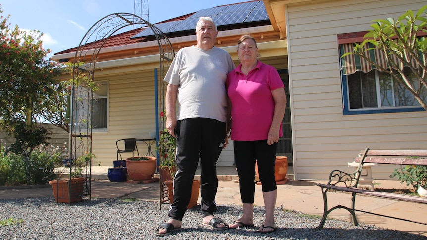 An older couple stand in the front garden of their home, which has solar panels on the roof