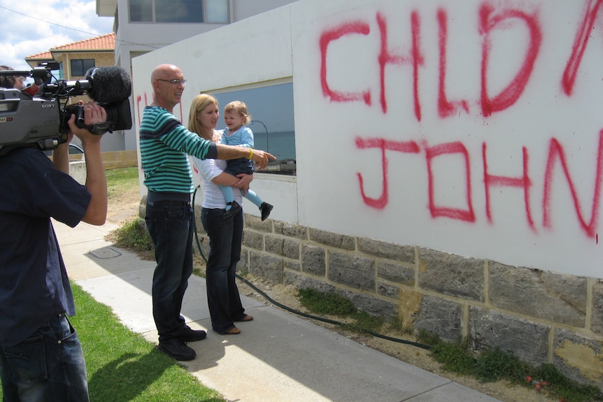 Three people stand in front a wall with graffiti sprayed on it and point