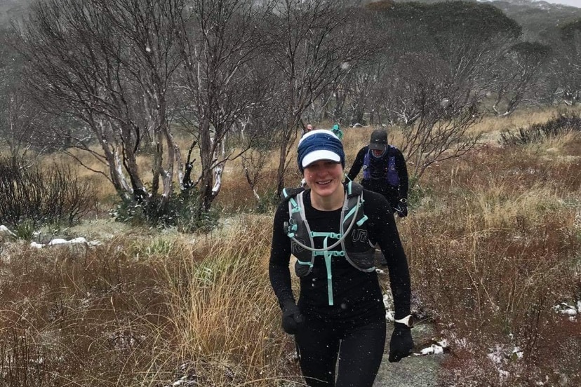 A woman in running gear stands on a mountain as snow falls lightly.