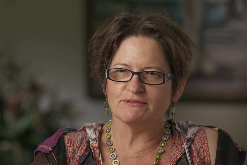 A woman wearing glasses and beaded jewellery sits for an interview, looking past the camera