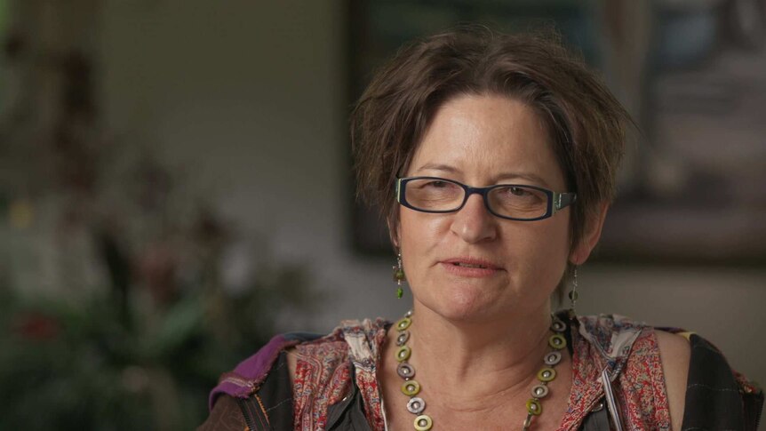 A woman wearing glasses and beaded jewellery sits for an interview, looking past the camera