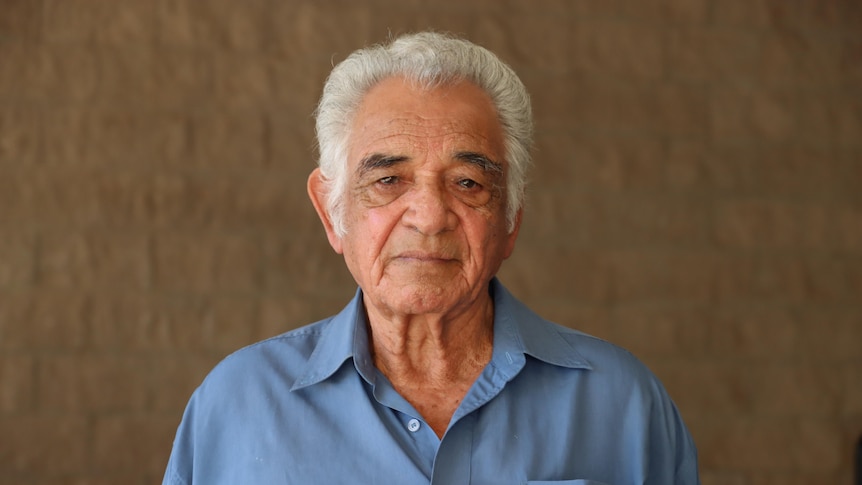 An Aboriginal man stands in front of a brick wall looking solemnly at the camera