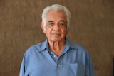 An Aboriginal man stands in front of a brick wall looking solemnly at the camera