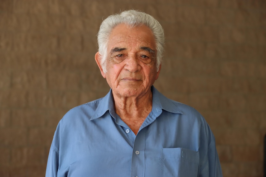 An Aboriginal man stands in front of a brick wall looking solemnly at the camera