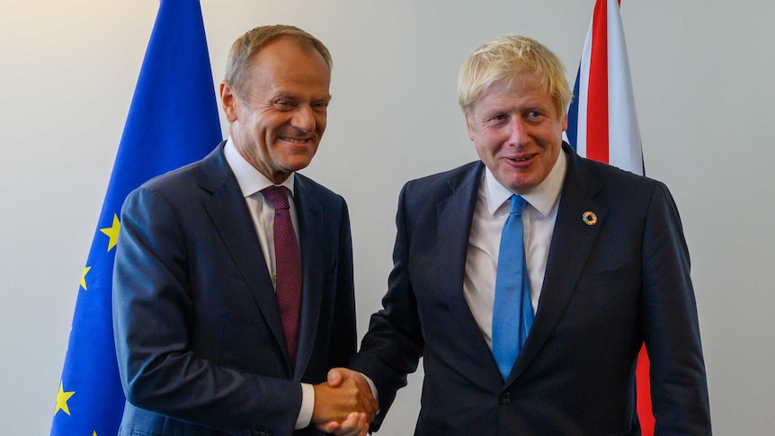 Donald Tusk shakes hands with Boris Johnson in front of EU and British flags.