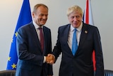 Donald Tusk shakes hands with Boris Johnson in front of EU and British flags.