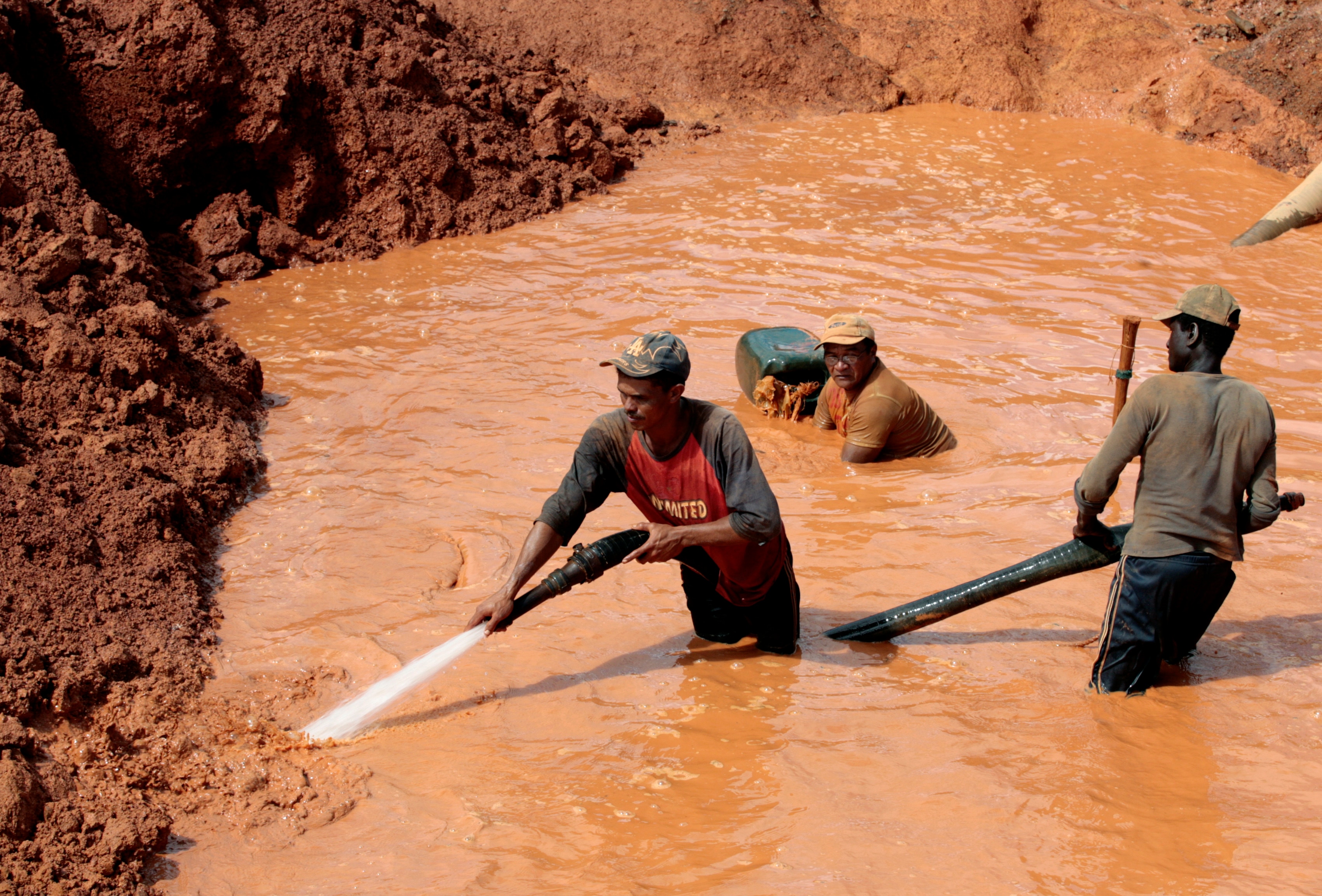 Three men standing in copper orange water, with two holding an active water hose, spraying mounds of red dirt.