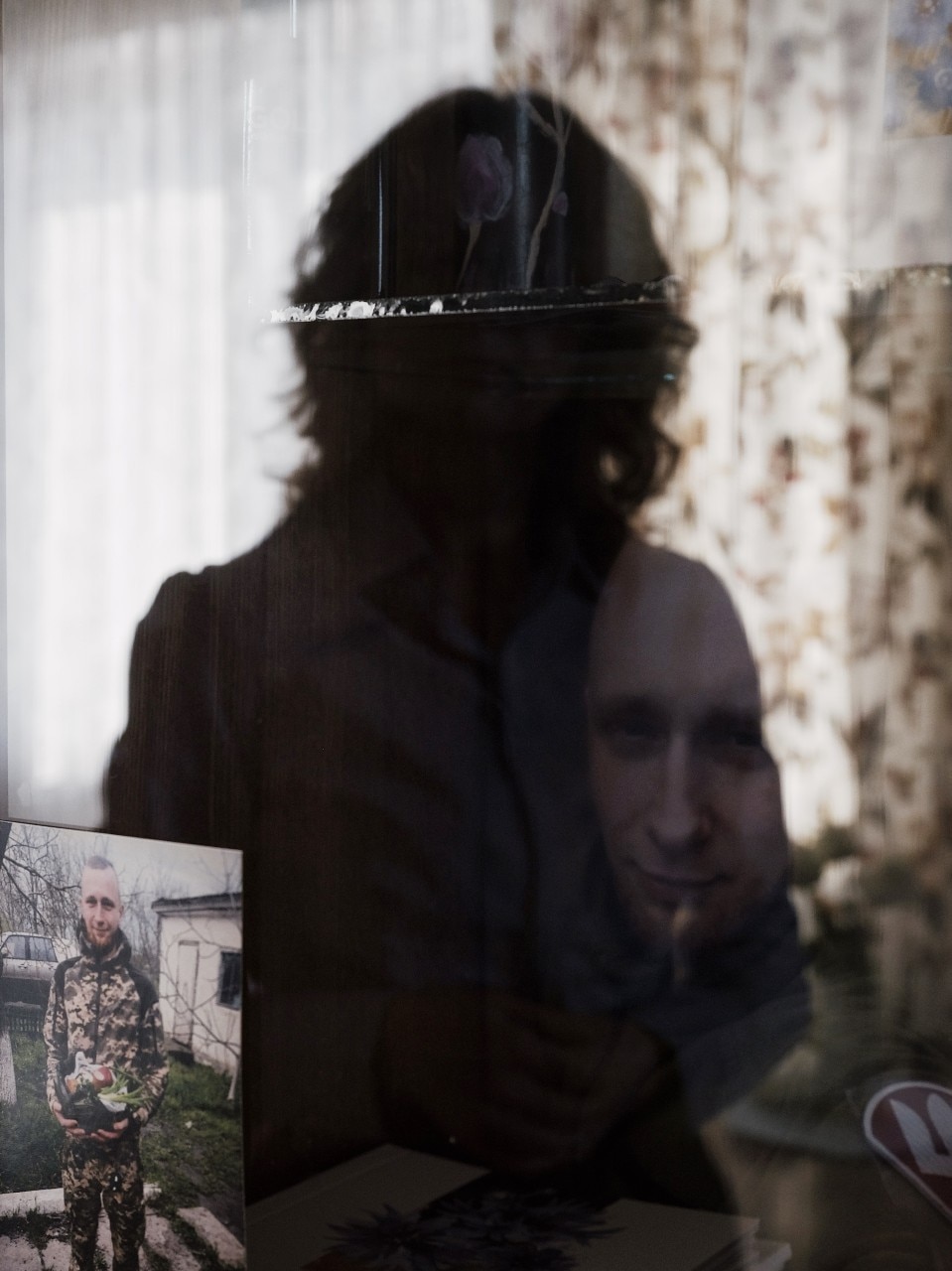 The silhouette of a woman is visible in a window, where a picture of a young man in military uniform is also visible.