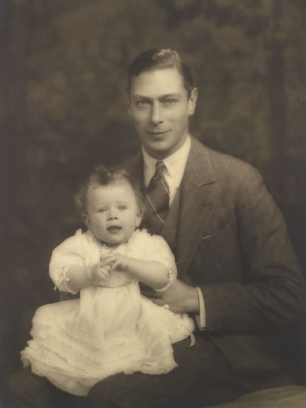King George VI holds a baby Queen Elizabeth on his knee in a black and white photograph.