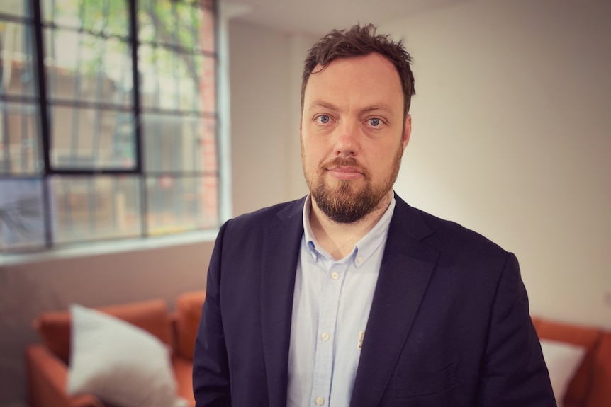 Brendan Coates, wearing a jacket, stands in an office looking at a camera.