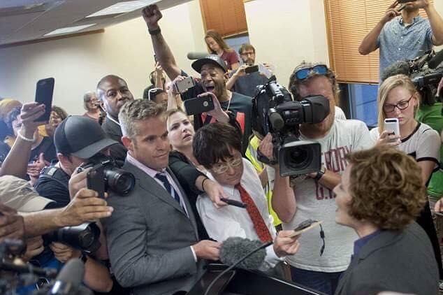 Duffy amongst crowd of journalists and camera operators interviewing a woman behind a podium.