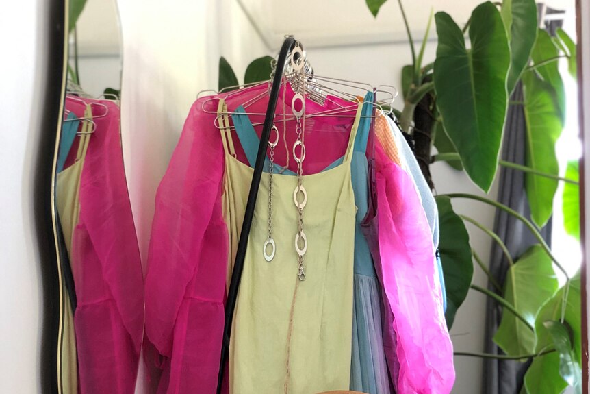 A mint and bright pink dress are seen hanging on an open clothes rail next to a mirror in a bedroom on a sunny day.