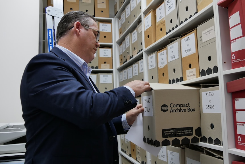 A man in a suit going through files in a room full of boxes. 