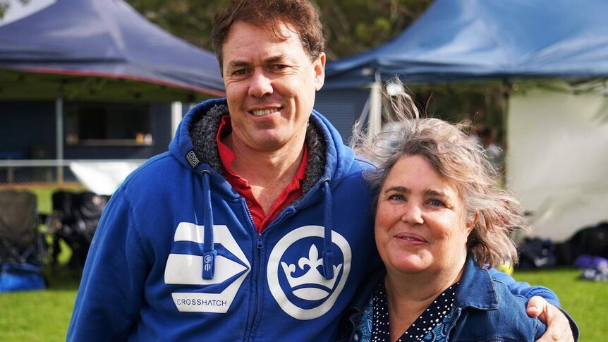 A man with a blue parker and a woman with a denim jacket stand smiling, arms around each other, in front of sport gazebos.
